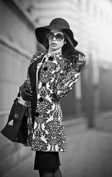 O femeie tânără atractivă într-o fotografie de modă de iarnă. Frumoasă fată tânără la modă în negru pozând pe bulevard. Elegant bruneta cu pălărie, ochelari de soare și geantă de mână în peisaj urban . — Fotografie, imagine de stoc