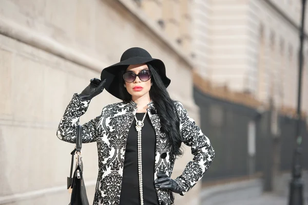 Attractive young woman in a winter fashion shot. Beautiful fashionable young girl in black posing on avenue. Elegant brunette with hat, sunglasses and handbag in urban scenery. — Stock Photo, Image