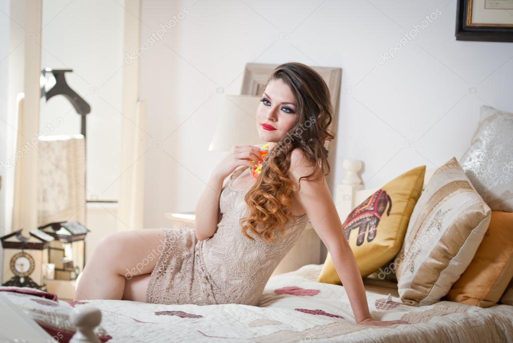 Young beautiful sexy woman in white short tight dress posing challenging indoor on vintage bed. Sensual long hair brunette in bedroom. Attractive female lying provocatively on bed full of pillows