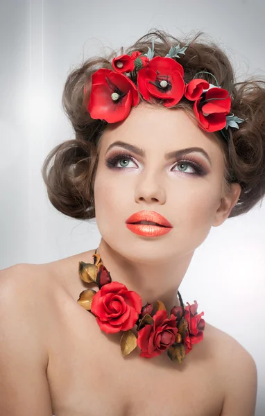 Retrato de una hermosa chica en el estudio con flores rojas en el pelo y rosas rojas alrededor del cuello. Mujer joven con maquillaje y flores brillantes como accesorios. Peinado creativo y maquillaje, estudio —  Fotos de Stock