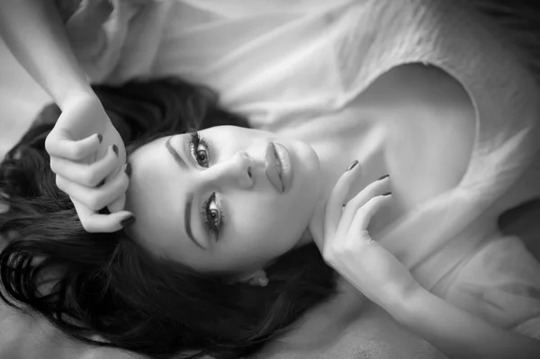 Beautiful sexy brunette young woman lying on bed. Sensual gorgeous perfect female posing provocatively, indoor shot. Attractive long hair girl with creative makeup and amazing eyes, black and white. — Stock Photo, Image