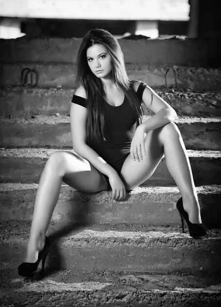 Fashionable pretty young woman with long legs sitting on old stone stairs. Beautiful long hair brunette on high heels shoes posing provocatively. Young model wearing black short dress, frontal view — Stock Photo, Image
