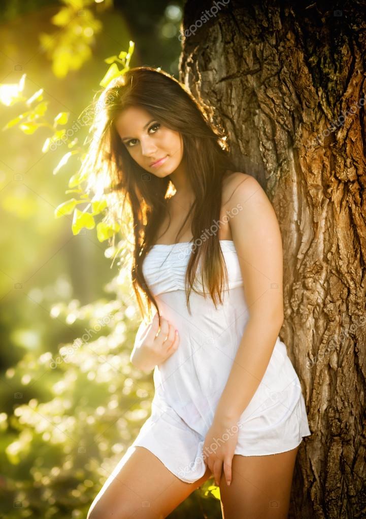 Attractive young woman in white short dress posing near a tree in a  