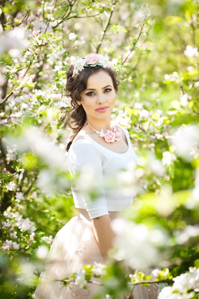 Retrato de una hermosa niña posando al aire libre con flores de los cerezos en flor durante un brillante día de primavera. Atractiva mujer morena con flores accesorios en huerto, tiro de primavera —  Fotos de Stock