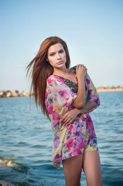 Joven chica de pelo rojo sexy en blusa multicolor posando en la playa. Sensual mujer atractiva con el pelo largo, tiro de verano en la orilla del mar. Cuerpo perfecto femenino, concepto de vacaciones turquesa fondo de agua —  Fotos de Stock