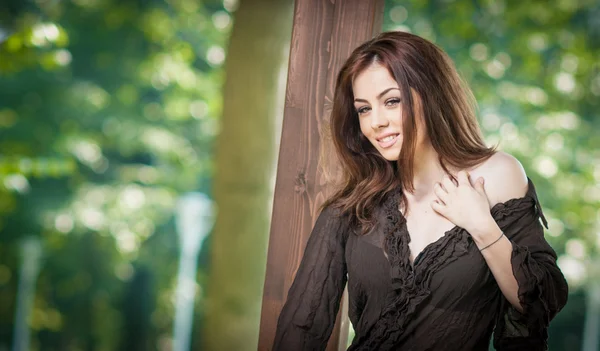 Bellissimo ritratto femminile con lunghi capelli castani all'aperto. Bruna naturale genuina con i capelli lunghi nel parco. Ritratto di una donna attraente con bellissimi occhi e camicetta nera vicino a un albero — Foto Stock