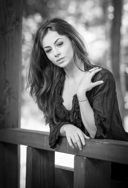 Hermoso retrato femenino con cabello castaño largo al aire libre. Genuina morena natural con magníficos ojos posando en el parque. Retrato de una mujer atractiva con vestido negro sentado en un pabellón de madera — Foto de Stock