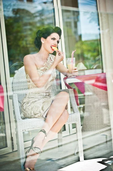Mujer joven atractiva de moda degustando una rodaja de limón en el restaurante, más allá de las ventanas. Hermosa morena posando en un elegante paisaje vintage con una copa de limonada. Concepto de foto a través de la ventana —  Fotos de Stock