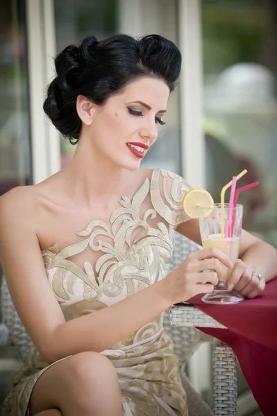 Mujer joven atractiva de moda en vestido de encaje sentado en el restaurante, más allá de las ventanas. Hermosa morena posando en un elegante paisaje vintage con una copa de limonada. Concepto de foto a través de la ventana —  Fotos de Stock