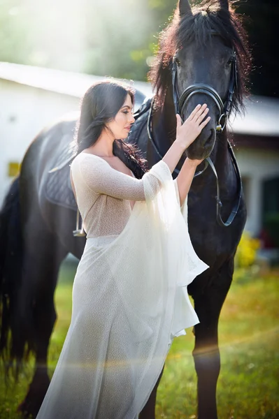 Fashionable lady with white bridal dress near brown horse. Beautiful young woman in a long dress posing with a friendly black horse. Attractive elegant female with horse, close-up photo — ストック写真