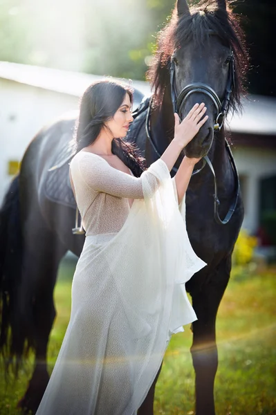 Señora de moda con vestido de novia blanco cerca de caballo marrón. Hermosa joven con un vestido largo posando con un amistoso caballo negro. Atractiva hembra elegante con caballo, foto de cerca —  Fotos de Stock