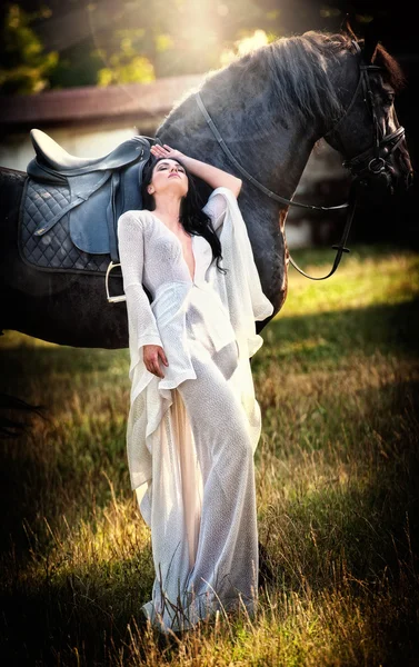 Fashionable lady with white bridal dress near brown horse in nature. Beautiful young woman in a long dress posing with a friendly black horse. Attractive elegant female with horse, sunny summer day — Stock Photo, Image