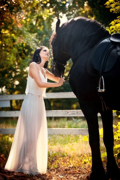 Модная дама в белом свадебном платье рядом с коричневой лошадью на природе. Красивая молодая женщина в длинном платье позирует с дружелюбной черной лошадью. Привлекательная элегантная женщина с лошадью, солнечный летний день — стоковое фото