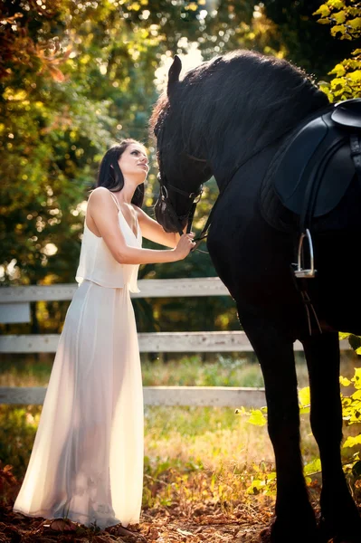 Donna alla moda con abito da sposa bianco vicino a cavallo marrone in natura. Bella giovane donna in un abito lungo in posa con un cavallo nero amichevole. Attraente elegante femmina con cavallo, soleggiata giornata estiva — Foto Stock