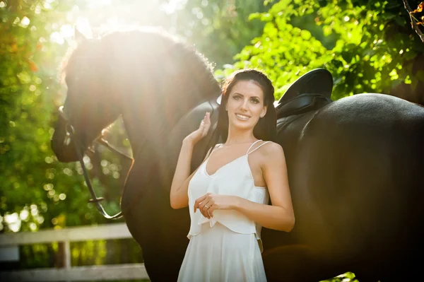 Модна леді з білим весільним платтям біля коричневого коня на природі. Красива молода жінка в довгій сукні позує з дружнім чорним конем. Приваблива елегантна жінка з конем, сонячний літній день — стокове фото