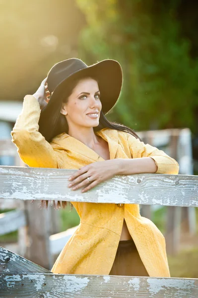 Hermosa chica morena con mirada campestre cerca de una vieja cerca de madera. Mujer atractiva con sombrero negro y abrigo amarillo, granjero de estilo americano. Cabello largo cabello oscuro femenino en estilo rústico —  Fotos de Stock