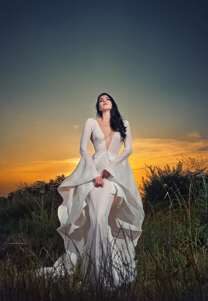 Fashionable beautiful young woman in white bridal long dress posing outdoor with dramatic sunset sky in background. Attractive long hair brunette girl with elegant luxurious dress, outdoors shot.