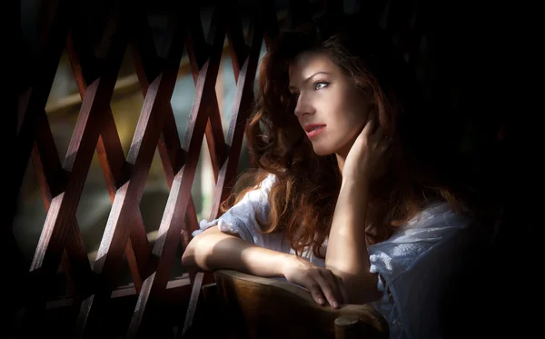 Charming young light brown hair woman looking through wooden grid. Sexy gorgeous young woman with long curly hair staying in rays of light. Beautiful portrait of a sensual woman with long hair — Stock Photo, Image