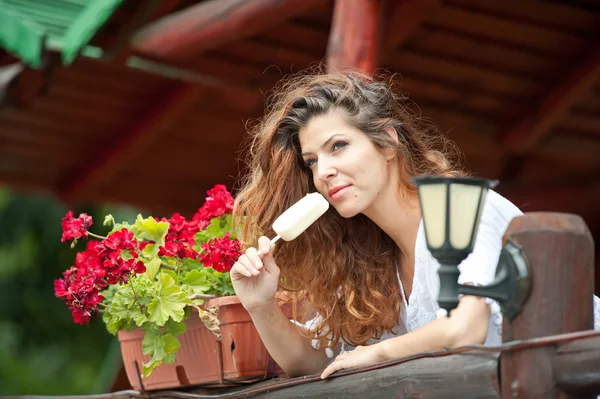 Beau portrait féminin avec de longs cheveux bruns mangeant de la crème glacée près d'un pot avec des fleurs rouges en plein air. Attrayant femme avec de beaux yeux souriant profiter d'une crème glacée dans une journée d'été, plan en plein air — Photo