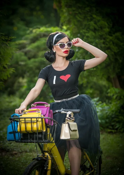 Hermosa chica con look retro vistiendo un traje negro divirtiéndose en el parque con bicicleta. Concepto de estilo de vida exterior. Paisaje vintage. Morena de moda con bicicleta y cesta con monederos de colores — Foto de Stock