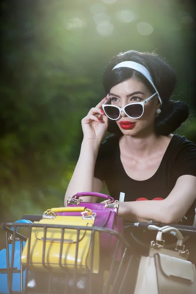Beautiful girl with retro look wearing a black outfit having fun in park with bicycle. Outdoor lifestyle concept. Vintage scenery. Fashionable brunette with bike and basket with colored purses — Stock Photo, Image