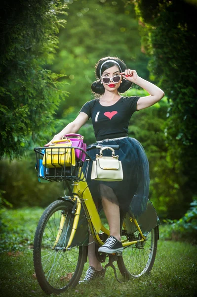 Beautiful girl with retro look wearing a black outfit having fun in park with bicycle. Outdoor lifestyle concept. Vintage scenery. Fashionable brunette with bike and basket with colored purses — Stock Photo, Image