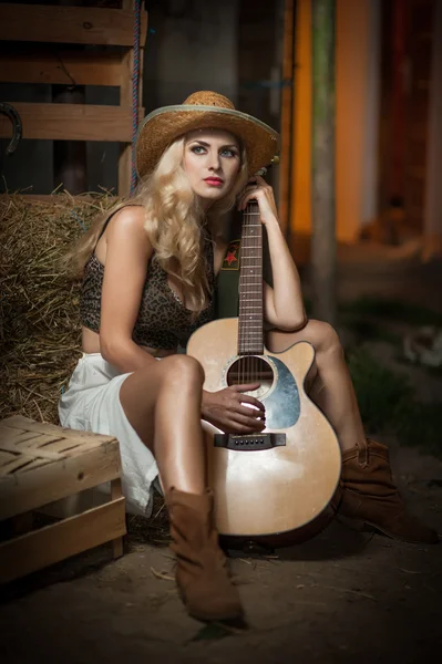 Attractive woman with country look, indoors shot, american country style. Blonde girl with straw cowboy hat and guitar. Fair hair female with boots posing with a guitar sitting on straw bale in barn — Stock Photo, Image