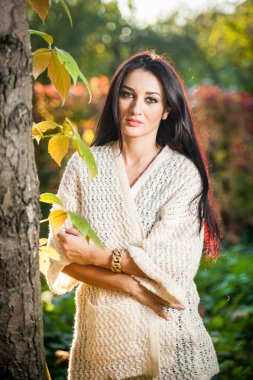 Beautiful woman in white posing in autumnal park. Young brunette woman spending time in autumn near a tree in forest. Long dark hair attractive woman smiling with faded leaves around her, outdoors