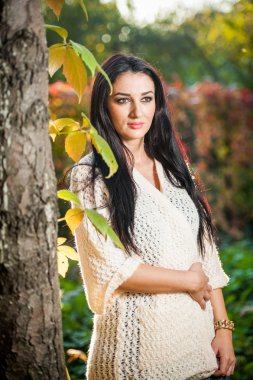 Beautiful woman in white posing in autumnal park. Young brunette woman spending time in autumn near a tree in forest. Long dark hair attractive woman smiling with faded leaves around her, outdoors