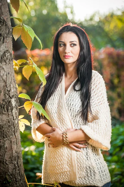 Beautiful woman in white posing in autumnal park. Young brunette woman spending time in autumn near a tree in forest. Long dark hair attractive woman smiling with faded leaves around her, outdoors — Stock Photo, Image