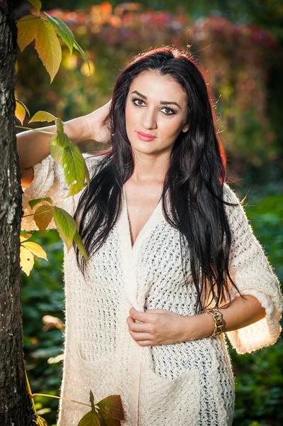 Beautiful woman in white posing in autumnal park. Young brunette woman spending time in autumn near a tree in forest. Long dark hair attractive woman smiling with faded leaves around her, outdoors — Stock Fotó