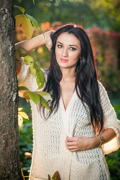 Beautiful woman in white posing in autumnal park. Young brunette woman spending time in autumn near a tree in forest. Long dark hair attractive woman smiling with faded leaves around her, outdoors — Stok fotoğraf