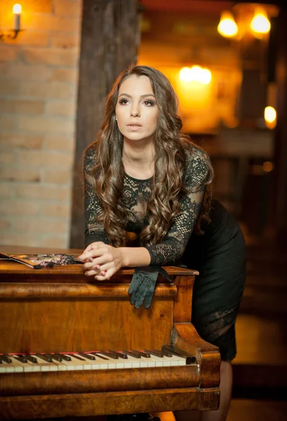 Young beautiful brunette woman in elegant black dress standing near a vintage piano. Sensual romantic lady with long dark hair in luxurious interior, daydreaming. Attractive girl posing indoors — Stock Photo, Image