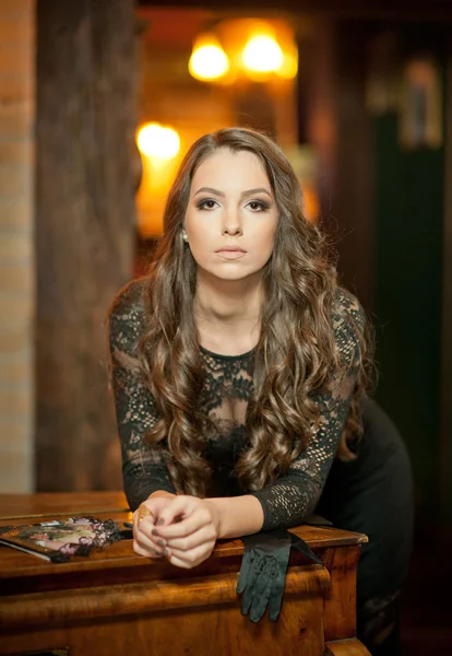 Young beautiful brunette woman in elegant black dress standing near a vintage piano. Sensual romantic lady with long dark hair in luxurious interior, daydreaming. Attractive girl posing indoors — Stock Photo, Image