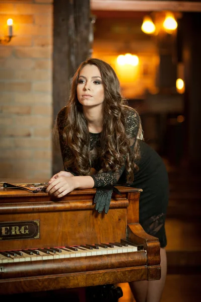 Mulher morena bonita nova no vestido preto elegante que está perto de um piano vintage. Sensual senhora romântica com longos cabelos escuros em interior luxuoso, sonhando acordado. Menina atraente posando dentro de casa — Fotografia de Stock