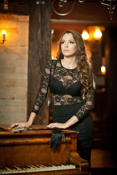 Young beautiful brunette woman in elegant black dress standing near a vintage piano. Sensual romantic lady with long dark hair in luxurious interior, daydreaming. Attractive girl posing indoors — Stock Photo, Image