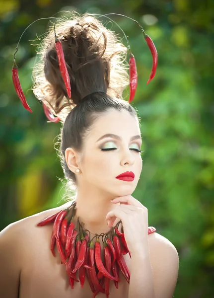 Mooie jonge vrouw portret met rode hete pikante pepers rond de hals en in haar, mannequin met creatieve voedsel plantaardige kapsel. Brunette meisje met rode chili paprika regeling outdoors. — Stockfoto