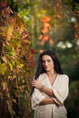 Beautiful woman in white posing in autumnal park. Young brunette woman spending time in autumn near a tree in forest. Long dark hair attractive woman smiling with faded leaves around her, outdoors