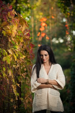 Beautiful woman in white posing in autumnal park. Young brunette woman spending time in autumn near a tree in forest. Long dark hair attractive woman smiling with faded leaves around her, outdoors