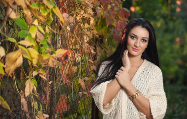Beautiful woman in white posing in autumnal park. Young brunette woman spending time in autumn near a tree in forest. Long dark hair attractive woman smiling with faded leaves around her, outdoors