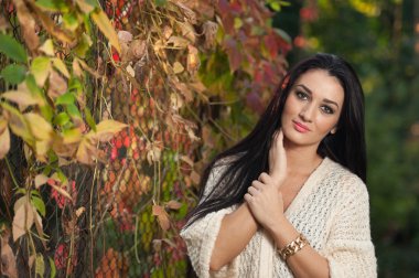Beautiful woman in white posing in autumnal park. Young brunette woman spending time in autumn near a tree in forest. Long dark hair attractive woman smiling with faded leaves around her, outdoors