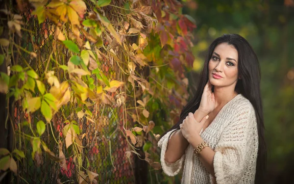 Beautiful woman in white posing in autumnal park. Young brunette woman spending time in autumn near a tree in forest. Long dark hair attractive woman smiling with faded leaves around her, outdoors — Stock Photo, Image
