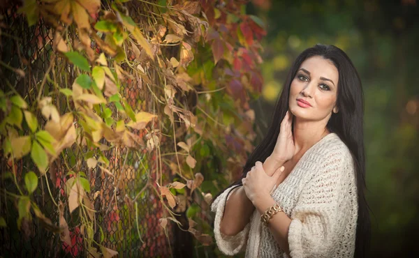 Beautiful woman in white posing in autumnal park. Young brunette woman spending time in autumn near a tree in forest. Long dark hair attractive woman smiling with faded leaves around her, outdoors — 图库照片