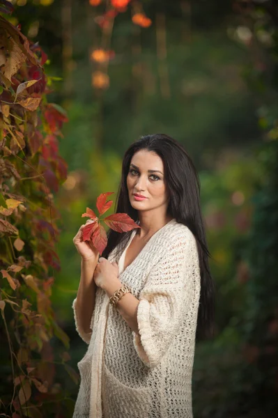 Beautiful woman in white posing in autumnal park. Young brunette woman spending time in autumn near a tree in forest. Long dark hair attractive woman smiling with faded leaves around her, outdoors — Stock fotografie