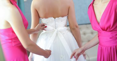 Two bride-maids in fuchsia dresses bustling the wedding dress. Bride-maids hands ribbon making on the back of bride white dress. Wedding preparation.