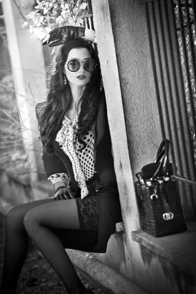 Attractive young woman with sunglasses in autumnal fashion shot. Beautiful lady in black and white outfit with short skirt sitting on wall. Elegant brunette, outdoors shot, black and white photo. — Stock Photo, Image