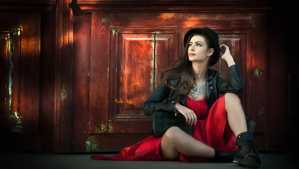 Joven mujer morena hermosa con vestido corto rojo y sombrero negro posando sensual en el paisaje vintage. Romántico misteriosa joven relajante sentado en el suelo contra la pared de madera vintage — Foto de Stock