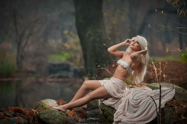 美丽的年轻女子坐在魔法森林的河流附近。性感的金发女郎，有着白色的衣服在秋天公园构成挑衅。童话的女孩看起来在秋天风景。浪漫的女人湖附近 — 图库照片