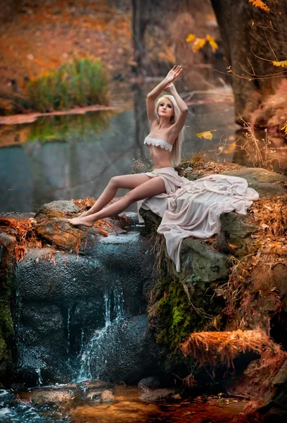 Lovely young lady sitting near river in enchanted woods. Sensual blonde with white clothes posing provocatively in autumnal park. Girl with fairy look in fall scenery. Romantic woman near lake — Stock Photo, Image