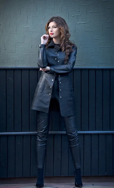 Charming young brunette woman in black leather outfit, coat and trousers, with dark gray wall on background. Sexy gorgeous young woman. Full length portrait of a sensual woman with long curly hair — Stock Photo, Image